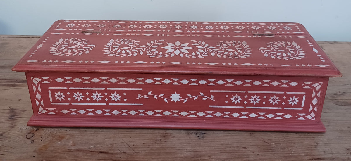 A rectangular red wooden storage box with white hand-stencilled geometric and floral patterns, resting on a rustic wooden surface against a plain light background.